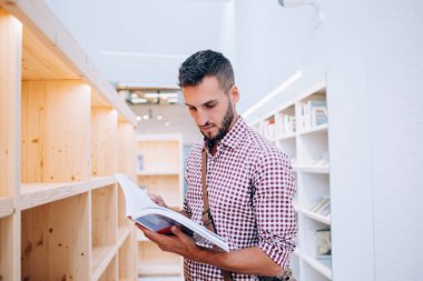 Kasaba kütüphanesinde kitaplarla dolu ahşap bir rafın önünde duran, çantasına günlük olarak kontrol edilmiş gömlek okuyan, zeki, genç ve sakallı bir erkek.