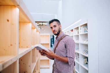 Şehir kütüphanesinde kitaplarla dolu ahşap bir rafın önünde duran, sıradan, sakallı, zeki, entellektüel bir genç adam.