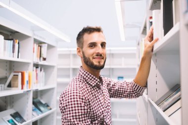 İyimser gülümseyen, gündelik giyinmiş, kitap arayan, kitapçıda fotoğraf makinesine bakan ve kitap okuyan etnik bir adamın yan görünüşü.