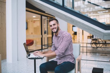 İyimser, mutlu, gündelik giyinen, iş yerinde kameraya bakarak işe başlamak için dizüstü bilgisayar kullanan bir etnik erkek.