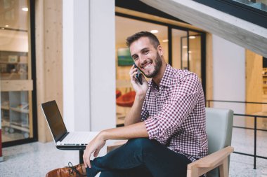 Keyifli gülümseyen, sakallı, normal tişörtlü, cep telefonuyla konuşan yaratıcı iş yerinde dizüstü bilgisayarlı genç bir erkek.