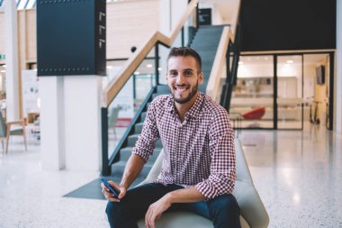 Keyifli gülümseyen, sakallı, normal tişörtlü, kot pantolonlu, cep telefonlu, yaratıcı iş yerinde sandalyede oturan, arka planda merdivenleri olan genç bir erkek.