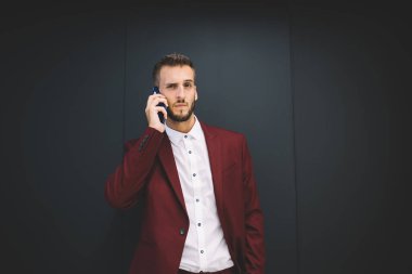 Odaklanmış, beyaz gömlekli, siyah düğmeli, kırmızı ceketli, gri arka planda cep telefonundan arayan ciddi, sakallı bir adam.