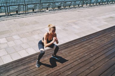 Spor giyimli ciddi bir kadın spor yapıyor güneşli bir günde gezinti güvertesindeki tahta merdivenlerde çömeliyor.