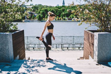 Siyah şık etek giyen konsantre kadın jimnastikçinin bir bacağını tutarken ve güneşli bir günde güzel rıhtımda uzanırken ayakta duruşu. 