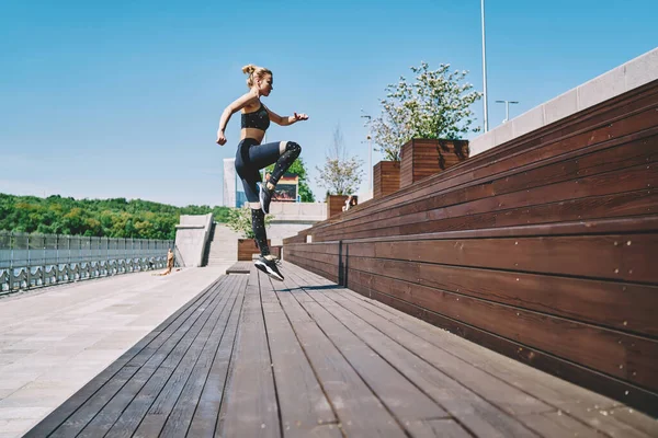 From below side view of active slim motivated female athlete jumping up with high knee lifting on wooden steps of city promenade in summer day