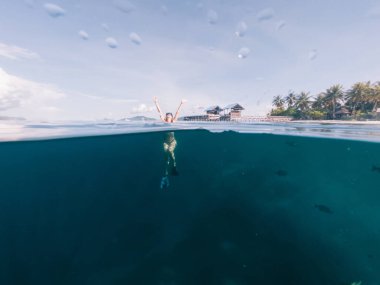 Gözlüklü, yüzgeçli, kırmızı bikinili genç bir sarışın yüzüyor ve bulutların ve rıhtımın arka planında ellerini kaldırarak yukarı bakıyor.