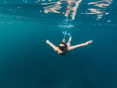 Kırmızı mayolu, yüzgeçli ve gözlüklü kahverengi saçlı genç bir kadın kameraya bakarken denizin altında yüzüyor.