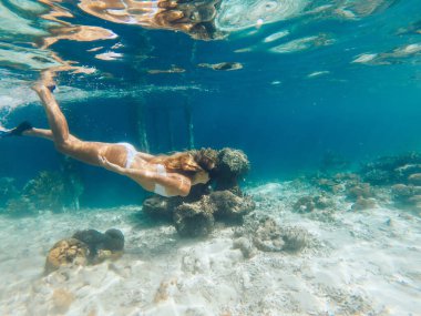 Beyaz bikinili, yüzgeçli ve gözlüklü genç sarışının, mercan resifi ve iskelenin yakınlarında denizin altında yüzüşünün yan görüntüsü.