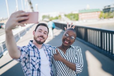 Günlük kıyafetlerle gülümseyen genç adam telefonda selfie çekiyor kısa saçlı siyahi kadınlara sarılıyor başparmağını kaldırıyor ve köprüde yürüyor. 