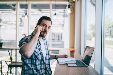 Odaklanmış serbest çalışan, mavi ekose gömlekli, parlak modern kafede oturan, telefonla konuşan, kameraya odaklanmış, günlük planlamacıda bilgisayar notları yazan ve güpegündüz kahve içen bir adam. 