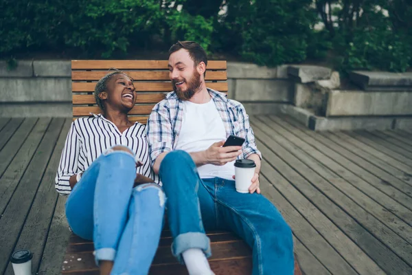 大人の多人種カップルがカジュアルな服を着て 木製のベンチに座って路上でスマートフォンを使用している間にお互いを見て笑いながら飲み物を取ります — ストック写真