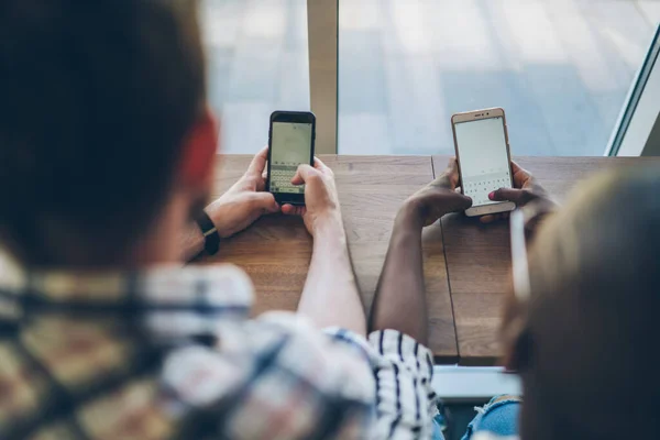 Crop from above view of multiracial people hands texting on smartphones and chatting in social media while sitting at table by large window