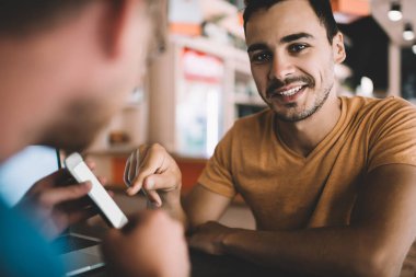 Akıllı telefonu işaret eden neşeli beyaz adamın portresi kafeteryada arkadaşlarıyla oturup sohbet eden, mutlu erkek blogcu hipster çocuk Colagu ile konuşmaktan memnun.