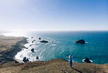 Dişi gezgin, seyahatte deniz manzarasının fotoğrafını çekiyor. Gezintiye çıkıyor, gezintiye çıkıyor. Okyanus kıyısındaki kayaların üzerinde durup fotoğraf çekiyor.