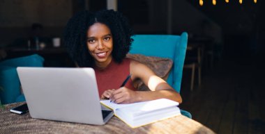 Siyah, neşeli, Afro saçlı, günlük kıyafetleri olan, not defterine not defteri yazan, kafede çalışmak için dizüstü bilgisayar kullanan genç bir kadın.