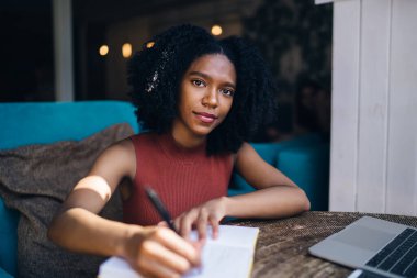 Gülümseyen kıvırcık saçlı etnik kadın not defterine not alıyor. Yazın şehirde kafede dizüstü bilgisayarla oturuyor.
