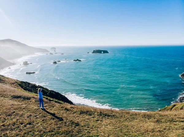 女旅行家站在岩石般的海洋海岸线上 在游手好闲的假期里欣赏着令人惊叹的风景和自然环境 而女旅行家则站在湾中观赏着从河岸到目的地的海景 — 图库照片