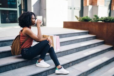 Afro-Amerikalı kadın, günlük giysiler içinde, kağıt poşetler içinde, paket kahve içiyor, yazın granit merdivenlerde oturuyor. 