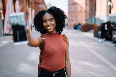 Heyecanlı, siyah kıvırcık saçlı, günlük giysiler içindeki genç bir kadın modern cep telefonunu gösteriyor. Şehrin caddesinde yürüyüp kameralara bakıyor.