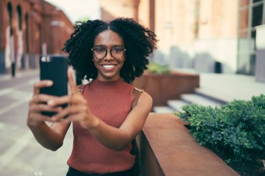 Afro saç stili ve gözlüklü neşeli siyah kadın akıllı telefon kullanıyor. Ünlü şehrin sokaklarında kameralara bakarak selfie çekiyor.