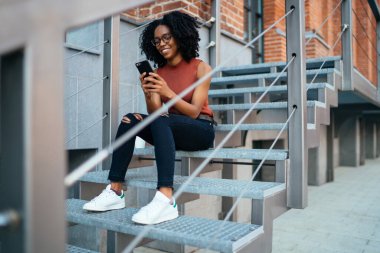 Afro-Amerikan kıvırcık saçlı, günlük giysiler ve gözlükler içinde telefon kullanarak merdivenlerde oturan bir arkadaşla mesajlaşmanın keyfini çıkaran bir kadın.