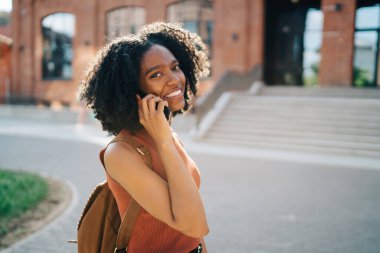 Sırt çantasıyla gülümseyen Afrikalı Amerikalı kız öğrencinin arka planda kameraya bakarken öğrenim ve öğretim binası girişi ile ilgili akıllı telefondan konuşmasının yan görüntüsü