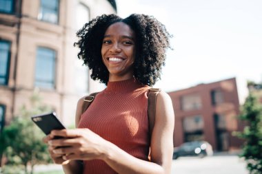 Gülümseyen, kıvırcık saçlı, kolsuz tişörtlü Afro-Amerikan kadın yazın sokakta cep telefonundan mesaj atıyor ve kameraya bakıyor.