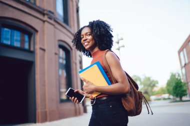 Mutlu, Afro-Amerikan kıvırcık saçlı, kolsuz gömlekli, akıllı telefon, kitap ve sırt çantasıyla gezen bir kadın.
