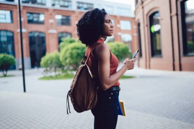 Afro saç stili olan Afro Amerikalı kız öğrencinin kampüste sokakta dururken cep telefonuyla mesajlaşmasının yan görüntüsü.