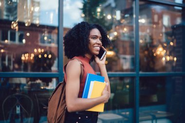 Afro saç stili ve notları akıllı telefondan konuşan, caddede cam binanın yanında duran neşeli Afro-Amerikan kız öğrencinin yan görüntüsü.