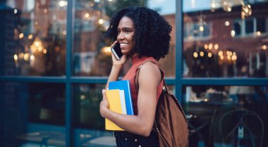 Arka planda cam duvarla cep telefonuyla konuşurken şık giyinmiş, gülümseyen Afro-Amerikan bayan öğrencinin yan görüntüsü