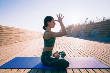 Mindere oturmuş, Lotus 'ta yoga yapan, arkada yanan ahşap güvertede sağlıklı bir hayat süren konsantre olmuş, sportif, ince kadın.