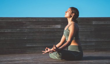 Nilüfer pozisyonunda, elleri dizlerinin üstünde, gözleri kapalı, gün doğumunda tahta patikada duran düşünceli, etnik bir kadın.