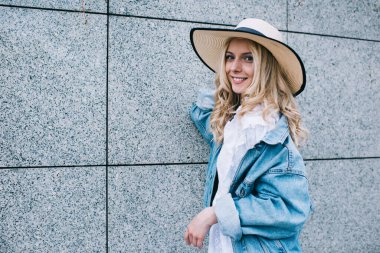 Yazlık şapkalı, kot ceketli, gülümseyen ve kameraya bakan çekici genç bir kadın.