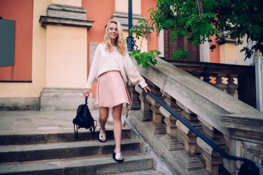Kendine güvenen, sarı saçlı, şık elbiseli çekici bir kadın sokak merdivenlerinde sırt çantasıyla yürüyor ve kameraya gülümsüyor. 