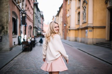 Eski kasabanın caddesinde yürüyen ve kameraya bakan neşeli, modern sarışın kadın. 