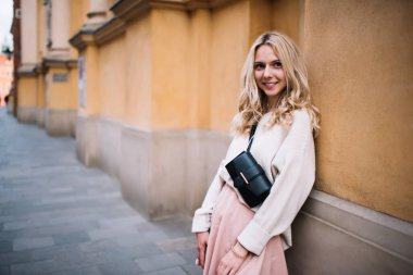 Moda kıyafetleri ve siyah kemer çantalı çekici genç sarışın eski şehrin duvarına yaslanıp kameraya bakıyor. 