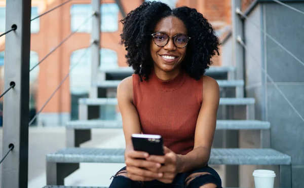 Black Glad Curly Haired Woman Casual Wear Glasses Surfing Smartphone — Stock Photo, Image