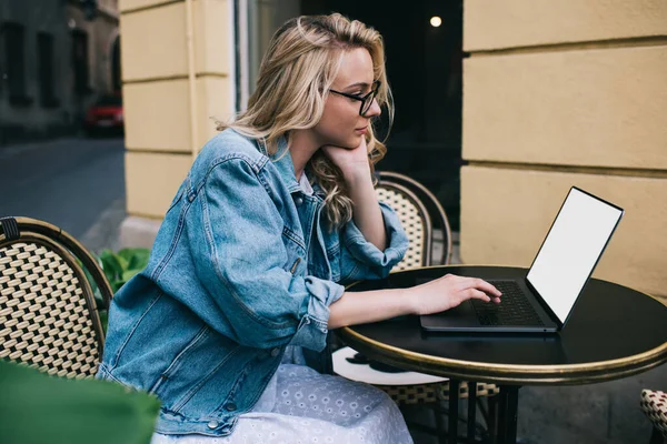 ストリートカフェのテーブル席に座っている間 空白の画面とノートパソコンを使用してデニムジャケットや眼鏡で集中若い女性のフリーランスの側面図 — ストック写真