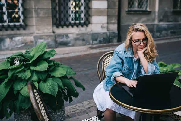 年轻的金发女人穿着时髦的斜纹棉布夹克和眼镜 坐在城市街上铺好的路边的咖啡桌旁 浏览着现代笔记本电脑 — 图库照片