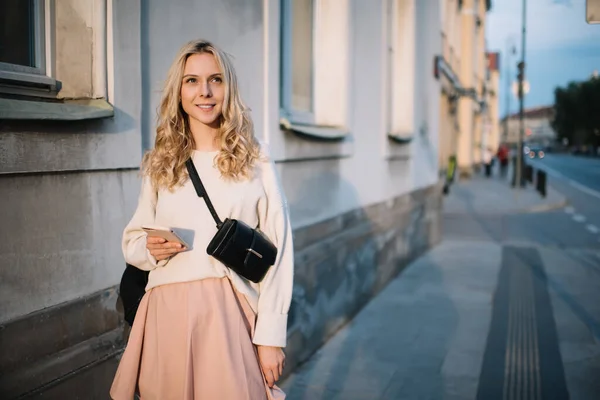 Young beautiful female in skirt and belt bag holding smartphone in hand and smiling away while having walk around town