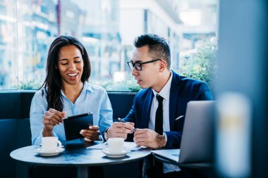 Modern kafede kahve molası sırasında masaya otururken ve dijital tabletle etkileşime girerken ekrana odaklanan ve gülen neşeli meslektaşlar.