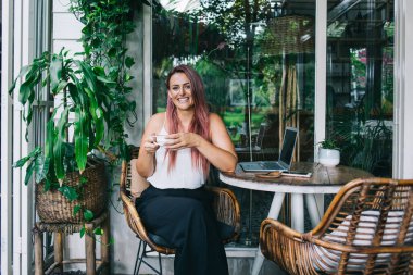 Rahat gülümseyen, kendine güvenen bir kadın açık teras kafesinde bacağının üzerinde oturuyor, dirseğini yuvarlak masaya dayamış, elinde bardak tutarken.