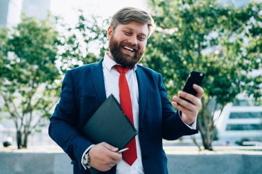 Resmi giyinmiş neşeli gülen adam, kırmızı kravatlı. Modern parkta cep telefonuyla sohbet ediyor.