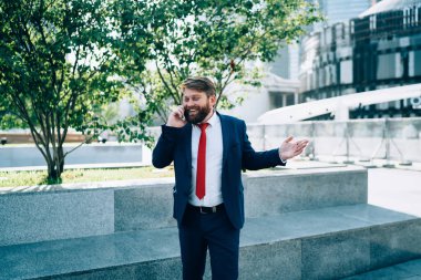 Mutlu, sakallı, şık takım elbiseli, kırmızı kravatlı, işe ara vermiş modern şehrin parkında akıllı telefondan arkadaşınla konuşan adam.