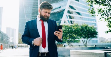 Koyu renk şık takım elbiseli ve kırmızı kravatlı dikkatli sakallı yönetici iş merkezinin karesinde cep telefonuna dikkatlice bakıyor.