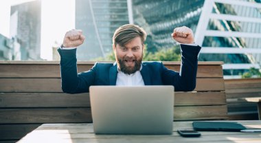 Mutlu sakallı iş adamı resmi olarak elleri kaldırmış başarılı bir açılışı kutlayan dizüstü bilgisayara bakıyor modern şehrin parkında masada oturuyor.