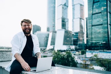 Modern dizüstü bilgisayarda yetenekli samimi hipster bir adamın portresi açık havada çalışan mutlu beyaz bir adam şehir ortamında portatif netbook 'la oturmuş ve neşeyle gülümsüyor.