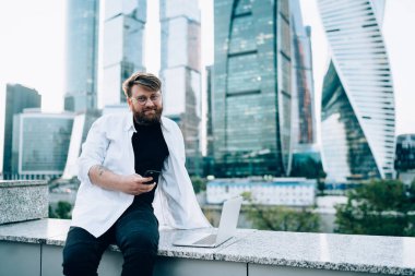 Görsel düzeltme için optik gözlüklü beyaz erkek programcının portresi kameraya bakıyor akıllı telefon bildirimini kontrol ediyor ve bilgi araştırması yapmak için dizüstü bilgisayar kullanıyor